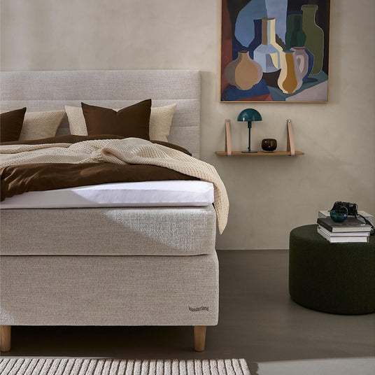 Beige polstret seng med brunt og cremefarvet sengetøj, mærket Wonderland, væghylde med grøn bordlampe og kunst over; mørkegrøn puff med bøger og kamera i et roligt soveværelse.