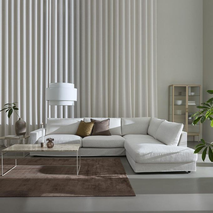 Lyst, minimalistisk stue med hvid hjørnesofa, marmorbord, brun tæppe, høje plisserede gardiner og grønne planter.