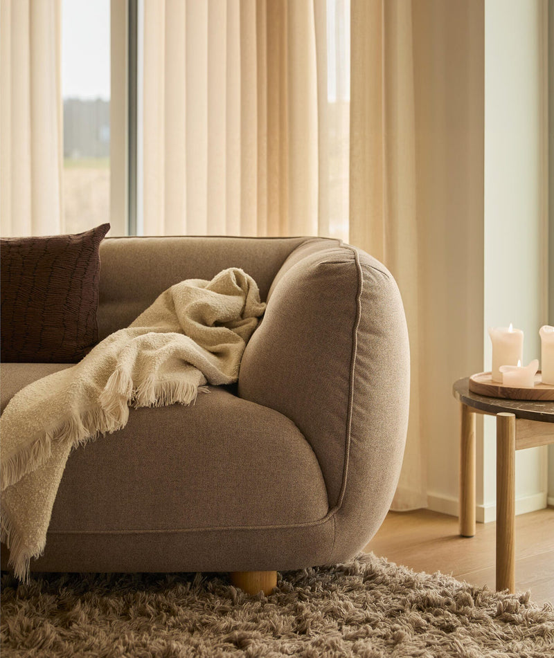 Nærbillede af et varmt, neutralt stuehjørne: brun sofa med cremefarvet plaid og pude, langluvet tæppe og lille træbord med tændte lys foran beige gardiner i dagslys.