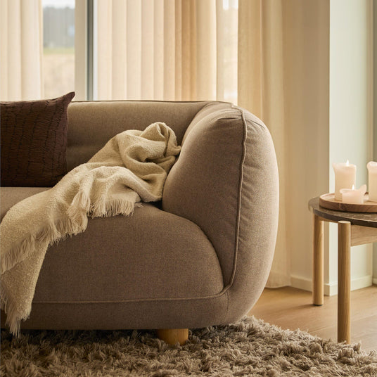 Nærbillede af et varmt, neutralt stuehjørne: brun sofa med cremefarvet plaid og pude, langluvet tæppe og lille træbord med tændte lys foran beige gardiner i dagslys.