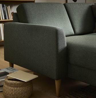 Nærbillede af grøn stofsofa med træben i stuen; fletkurv med notesbog og bogen A Modern Way to Live ved siden af, bogreol i baggrunden.