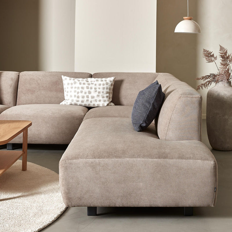 Beige hjørnesofa i stof med to pyntepuder, rundt lyst tæppe, sofabord i træ, pendellampe og stor vase med tørrede grene i en lys stue.