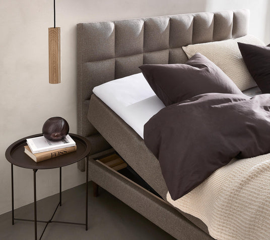 Neutralt soveværelse med polstret sengegavl, mørkebrunt sengetøj, beige strikket plaid og et rundt sidebord med en røgfarvet glasdekoration og to bøger, hvoraf den ene hedder FOSS; træpendel over bordet.