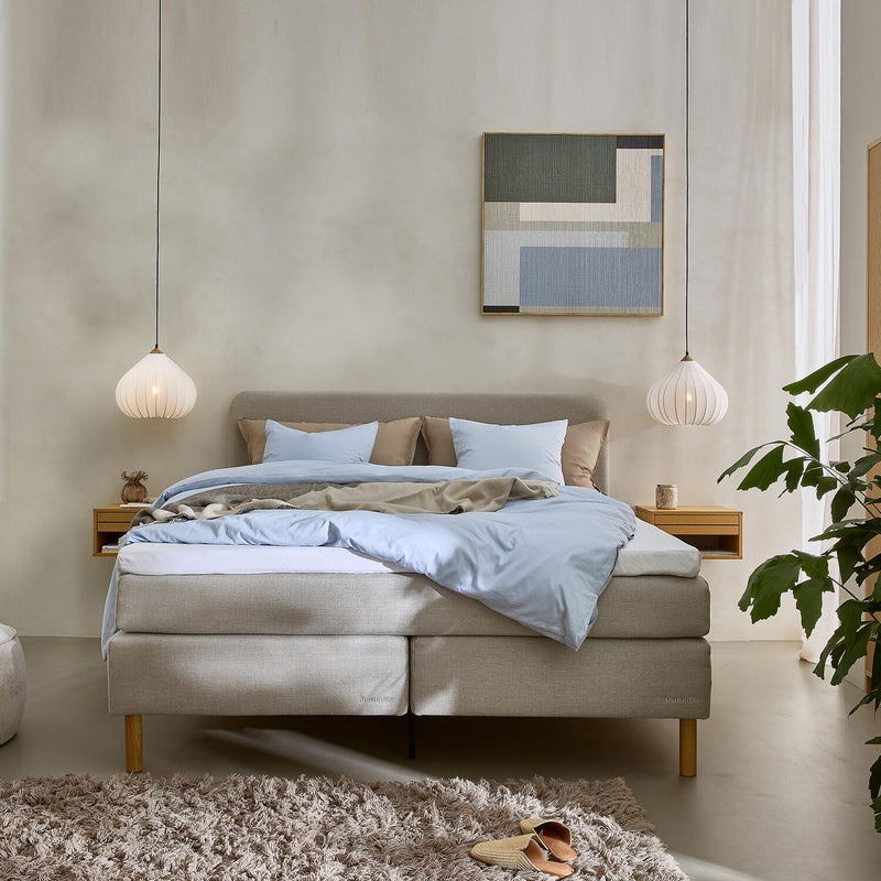 Lyst soveværelse med beige kontinentalseng redt med lyseblåt sengetøj, to hængende hvide pendellamper, abstrakt vægkunst, grønne planter og en blød beige ryatæppe med tøfler på gulvet.