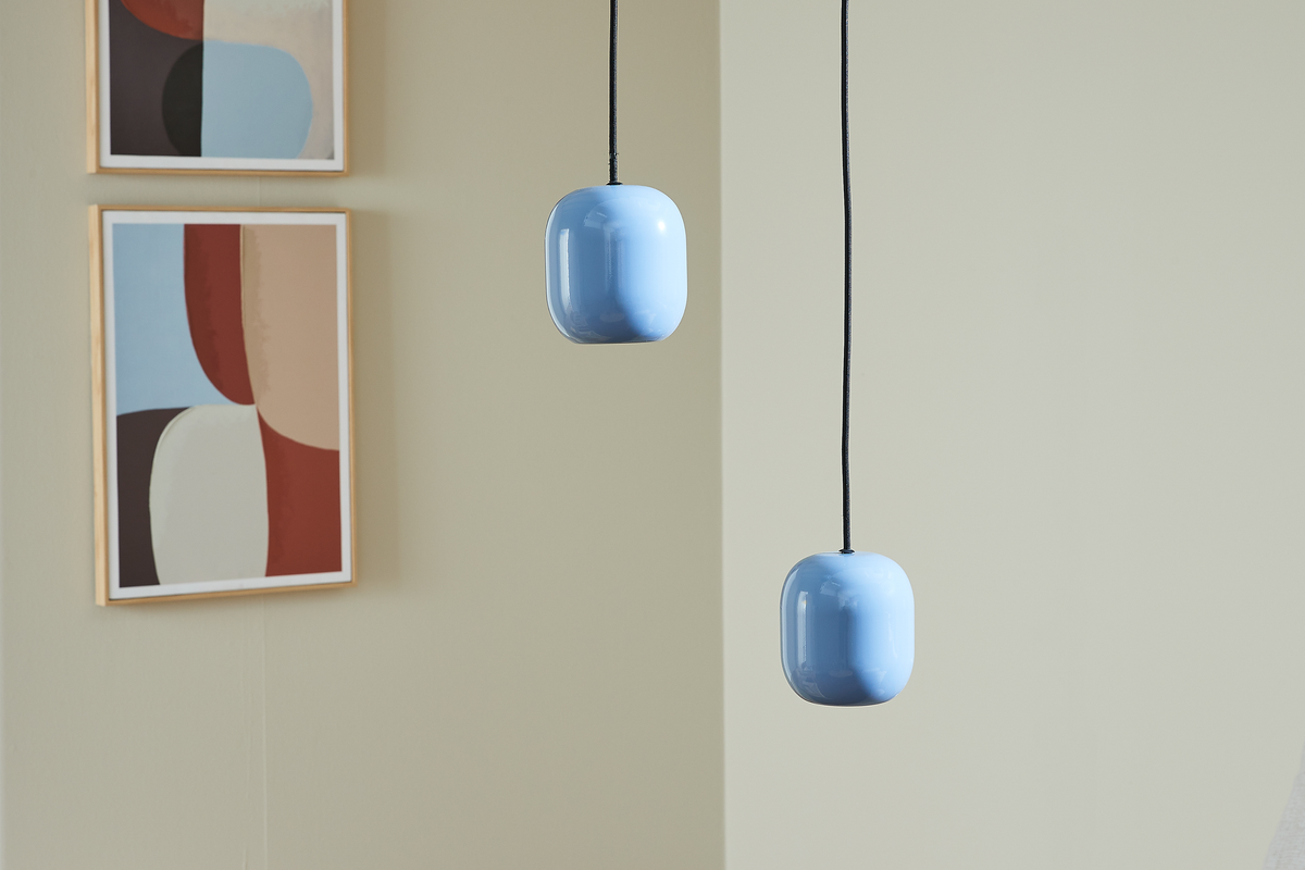 To lyseblå pendellamper med sorte ledninger foran en beige væg, med abstrakt vægkunst i trærammer i baggrunden.