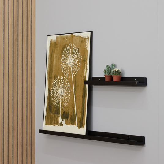 Sort billedhylde på væg med plakat i okker motiv og tre små kaktusser på en kortere hylde.