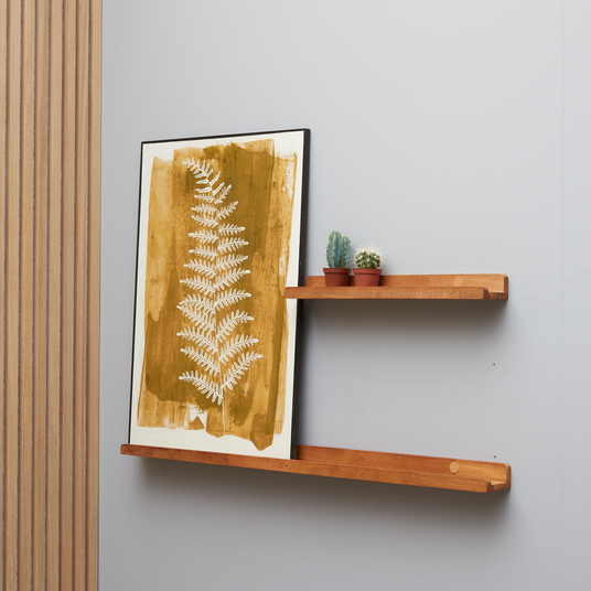 To vægmonterede træhylder med en sennepsgul botanisk plakat og to små kaktusser på en grå væg.