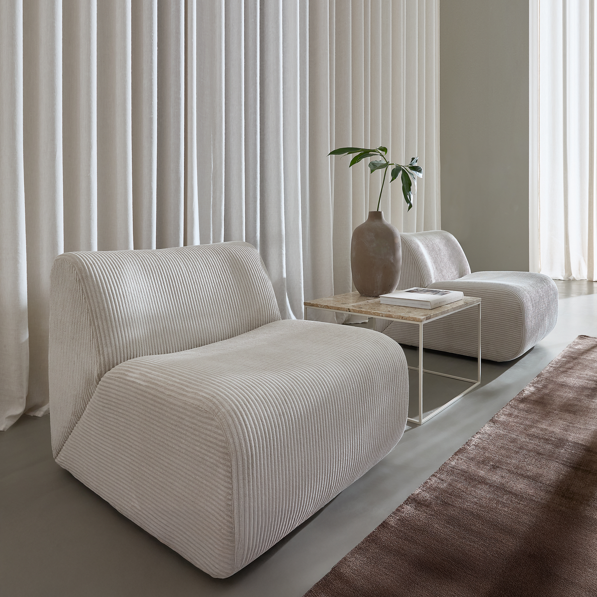 To lave loungestole i lys beige ribbet stof med et smalt bord med stenplade imellem, vase med grønne blade og brun tæppe foran samt lyse gulv-til-loft gardiner i blødt dagslys.