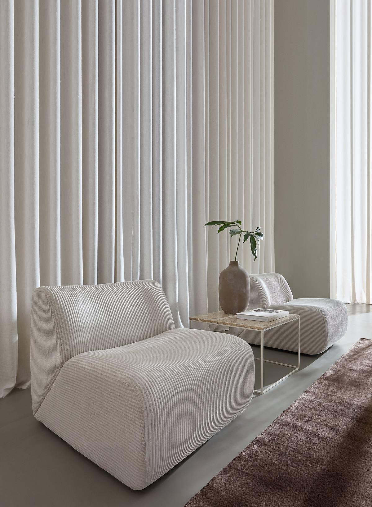 To lave beige loungestole i ribbet stof ved siden af et slankt sidebord med stenplade, keramisk vase med grøn kvist, gulv‑til‑loft‑gardiner og et brunt tæppe i en minimalistisk stue.