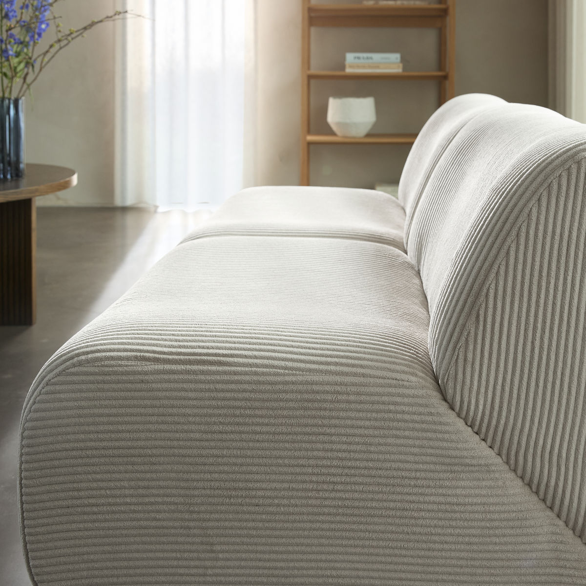 Nærbillede af beige riflede sofa i stue med reol, vase med blomster og blødt dagslys.
