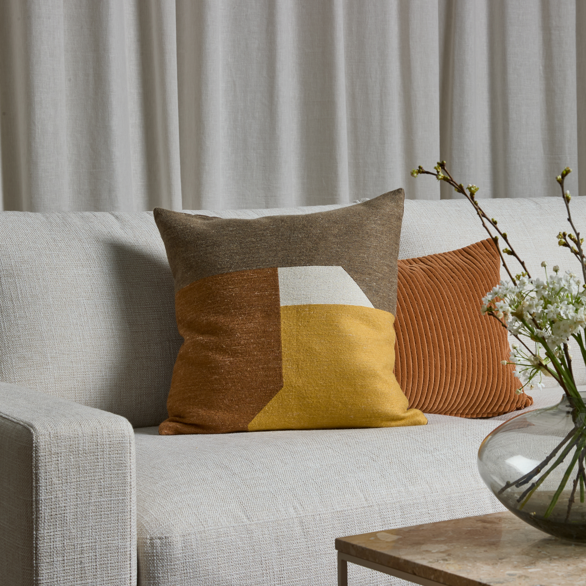 Lysegrå sofa med to pyntepuder – én med geometrisk mønster i sennep, brun og hvid og en rillet i rust – foran beige gardiner og en glasvase med hvide blomster på et stuebord i sten.