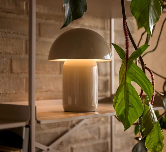 Svampeformet bordlampe i creme lyser varmt på en hylde ved siden af grønne planter.