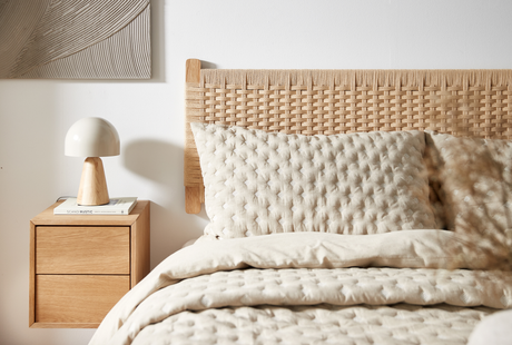 Lyst soveværelse med flettet sengegavl, beige quiltet sengetøj og et væghængt natbord med kuppelformet bordlampe oven på bogen Scandi Rustic.