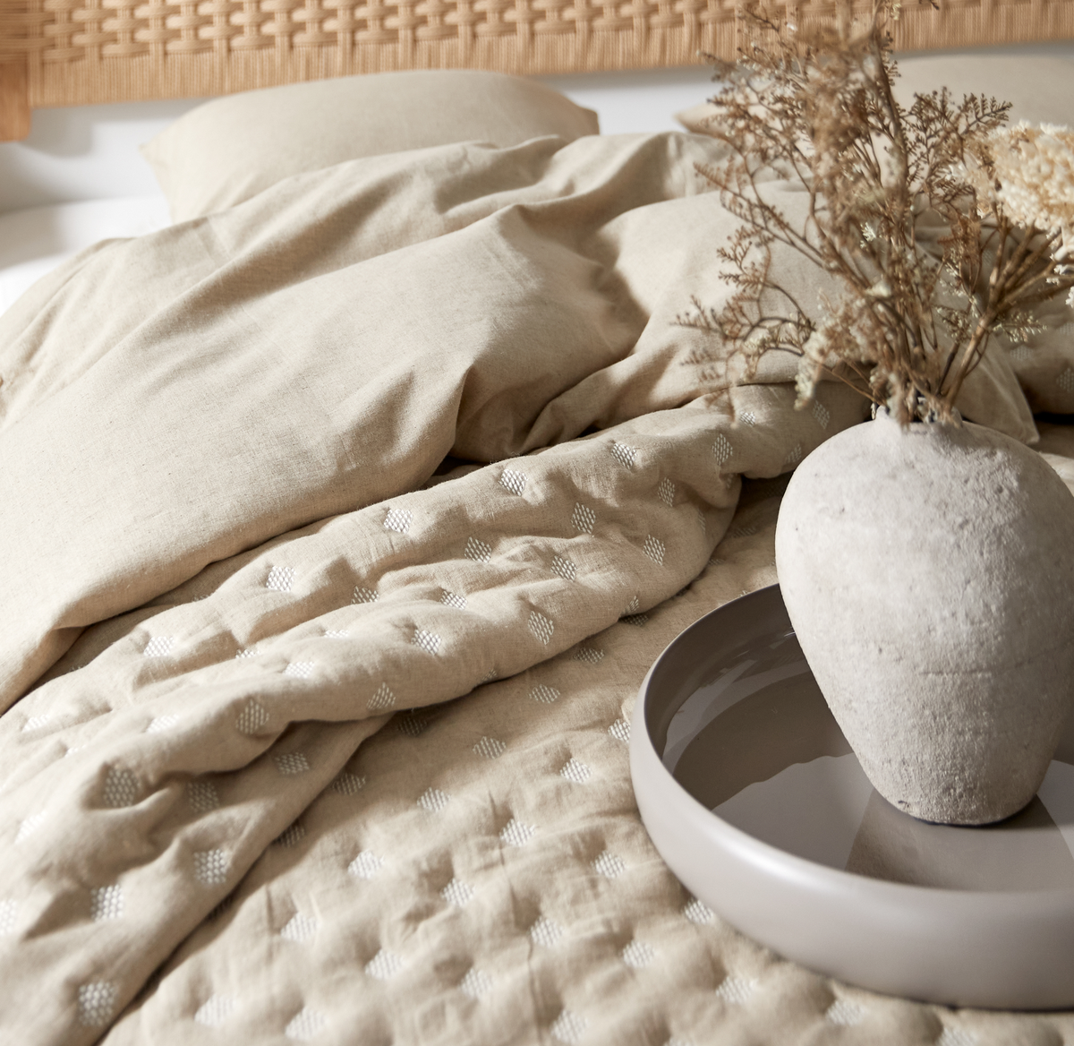 Beige sengetøj i linned-look med quiltet tæppe med små hvide prikker; keramikvase med tørrede blomster på rund bakke på sengen, flettet sengegærde i baggrunden.