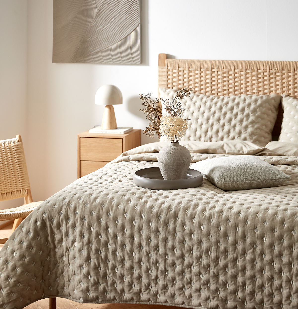 Beige soveværelse med quiltet sengetæppe og puder, fletgavel og natbord i træ; en grå bakke med vase og tørrede blomster står på sengen.