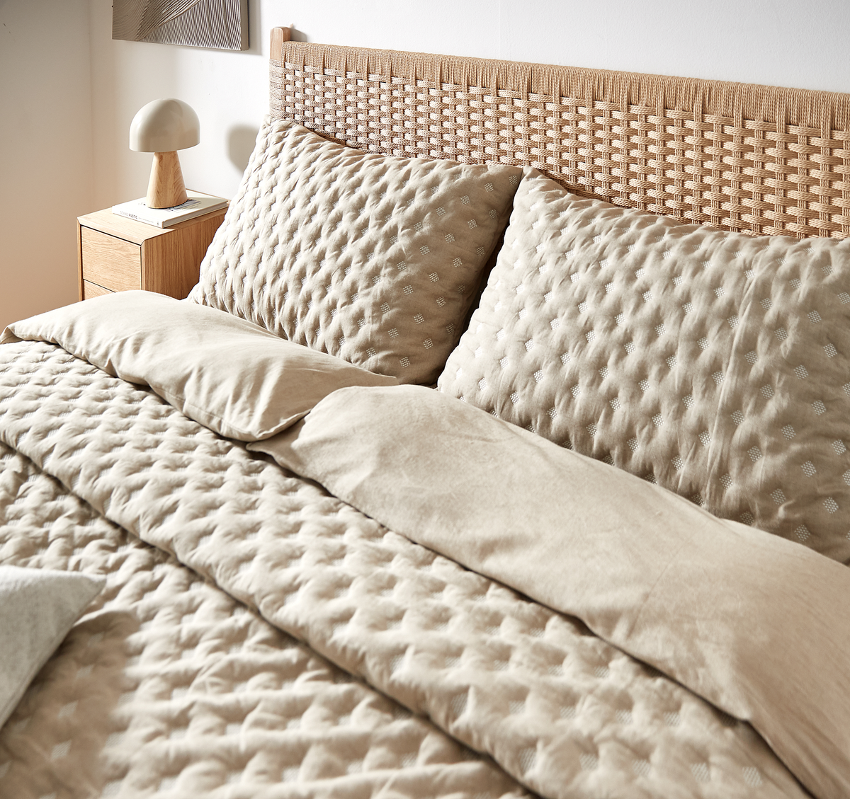 Beige quiltet sengetøj med store puder mod en rattan-sengegavl og natbord med lampe i blødt dagslys.