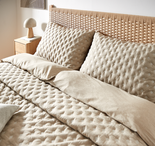Beige quiltet sengetøj med store puder mod en rattan-sengegavl og natbord med lampe i blødt dagslys.