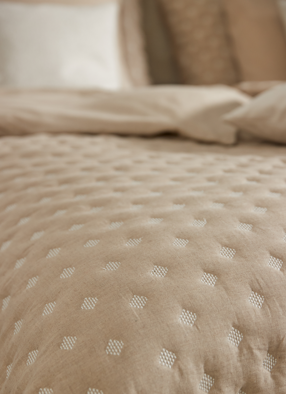 Nærbillede af beige, quiltet sengetekstil med små hvide diamantprikker; puder i neutrale toner i baggrunden.