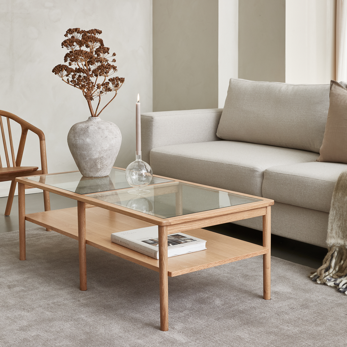 Lys stue med sofabord i eg med glasplade, vase med tørrede blomster, tændt lys i glas og en bog på nederste hylde foran en lysegrå sofa.