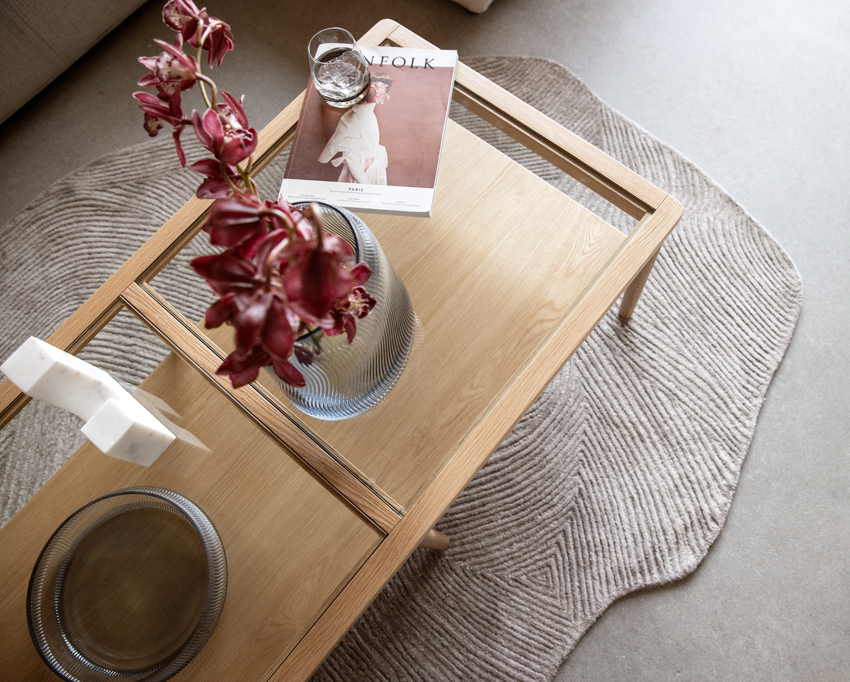 Lyst sofabord i træ med glasfelter, en vase med røde orkidéer, et glas med is og magasinet Kinfolk på et beige, tekstureret tæppe.