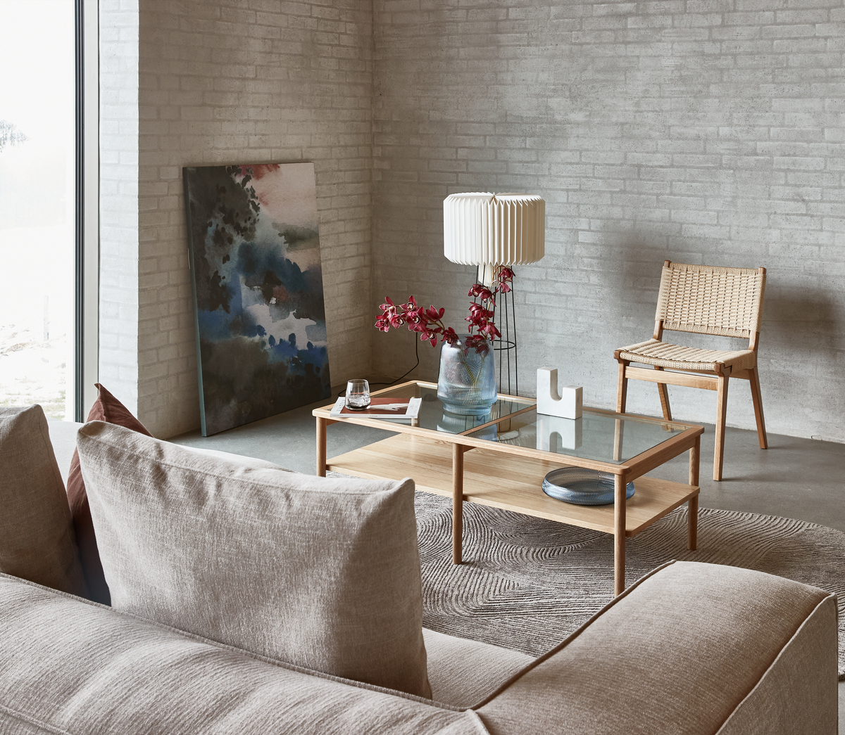 Lyst stue med beige sofa, glas-sofabord i træ, gulvlampe, fletstol og vase med røde blomster; maleri lænet op ad den lyse murstensvæg.