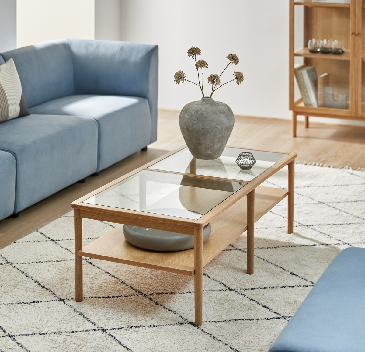 Lys stue med blå sofa, sofabord i træ med glasplade, vase med tørrede blomster og lyst tæppe med rudemønster.
