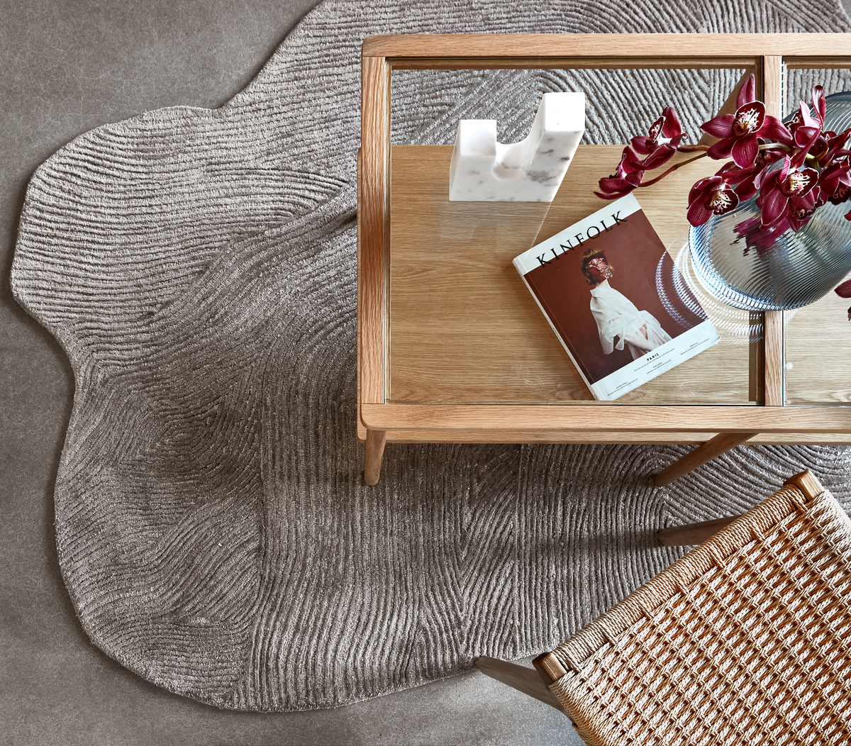Top-down af et sofa-/kaffebord i træ og glas på et tekstureret gråt tæppe, med et Kinfolk-magasin, en hvid marmordekoration, en riflet glasvase med mørkerøde orkidéer og en stol med flet sæde.