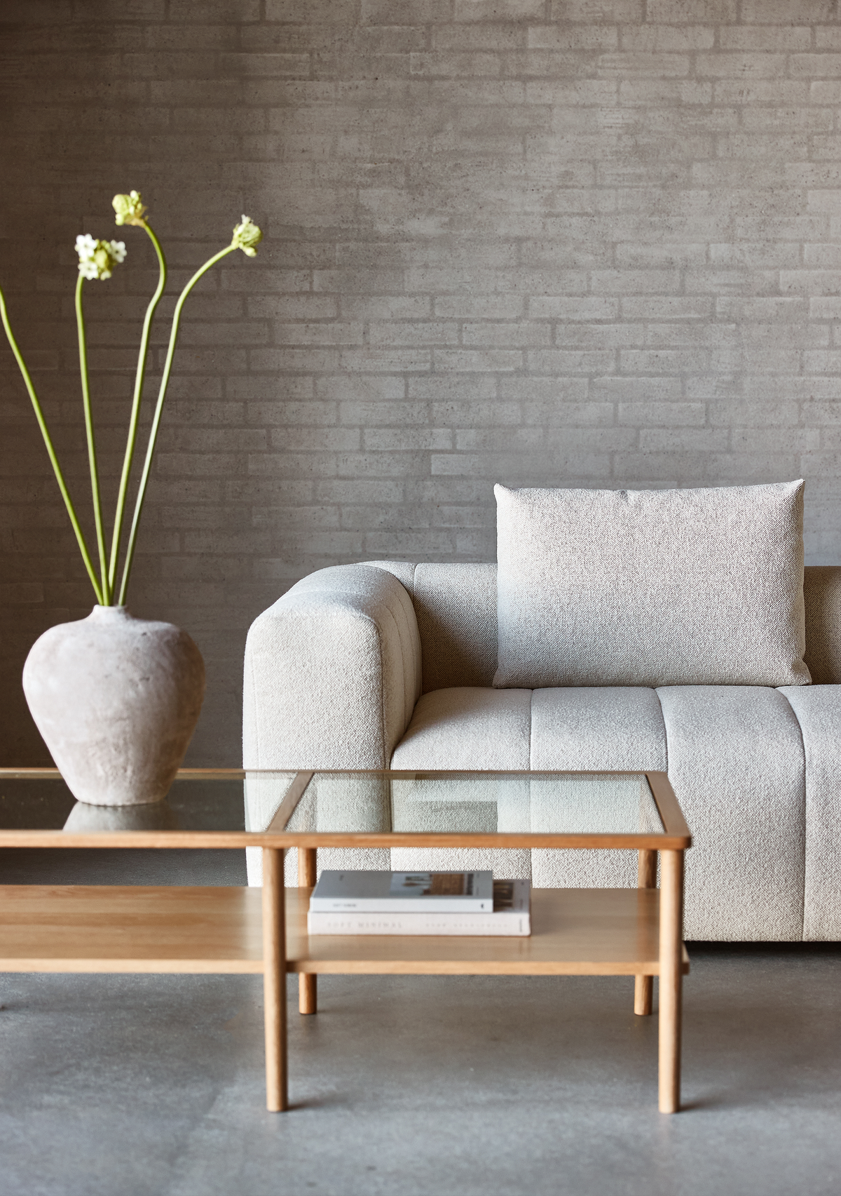 Beige bouclésofa med pude ved siden af et træ- og glas-sofabord med en sten-vase med lange blomsterstængler foran en grå murstensvæg.