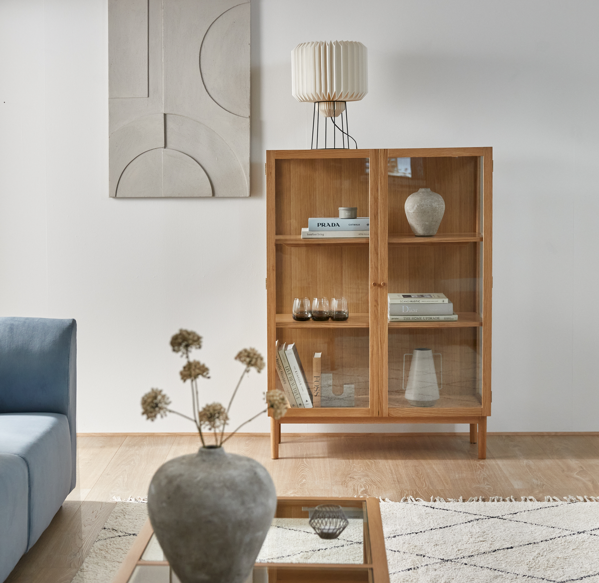 Lyst skandinavisk stue med vitrineskab i træ, bogtitlerne PRADA og DIOR på hylderne, blå sofa, vase på sofabord og plisseret bordlampe.
