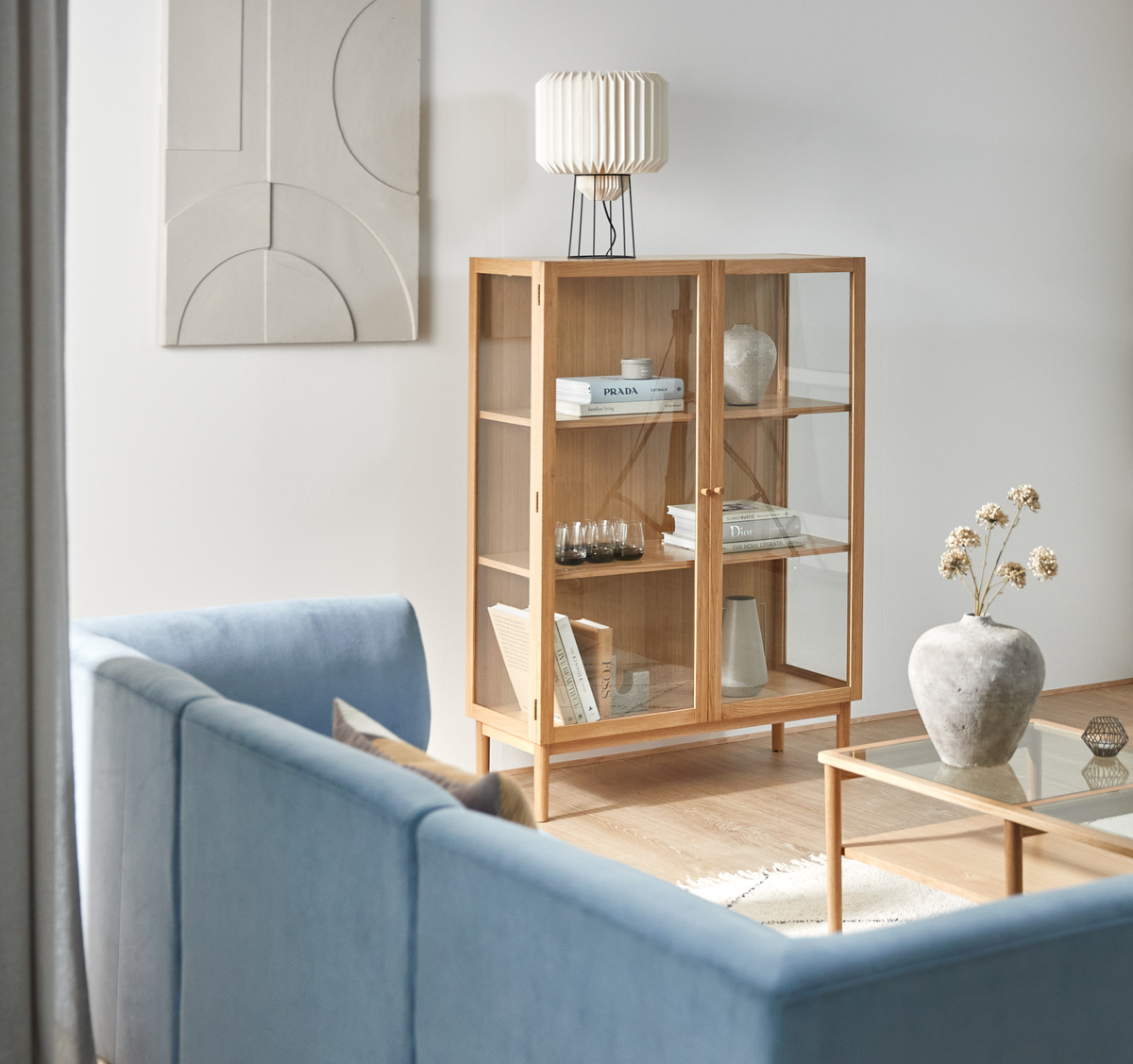 Lys stue med lyseblå sofa, glas- og træsofabord og vitrineskab i eg med bøgerne PRADA og Dior samt en plisseret bordlampe; minimalistisk dekoration og vægkunst.
