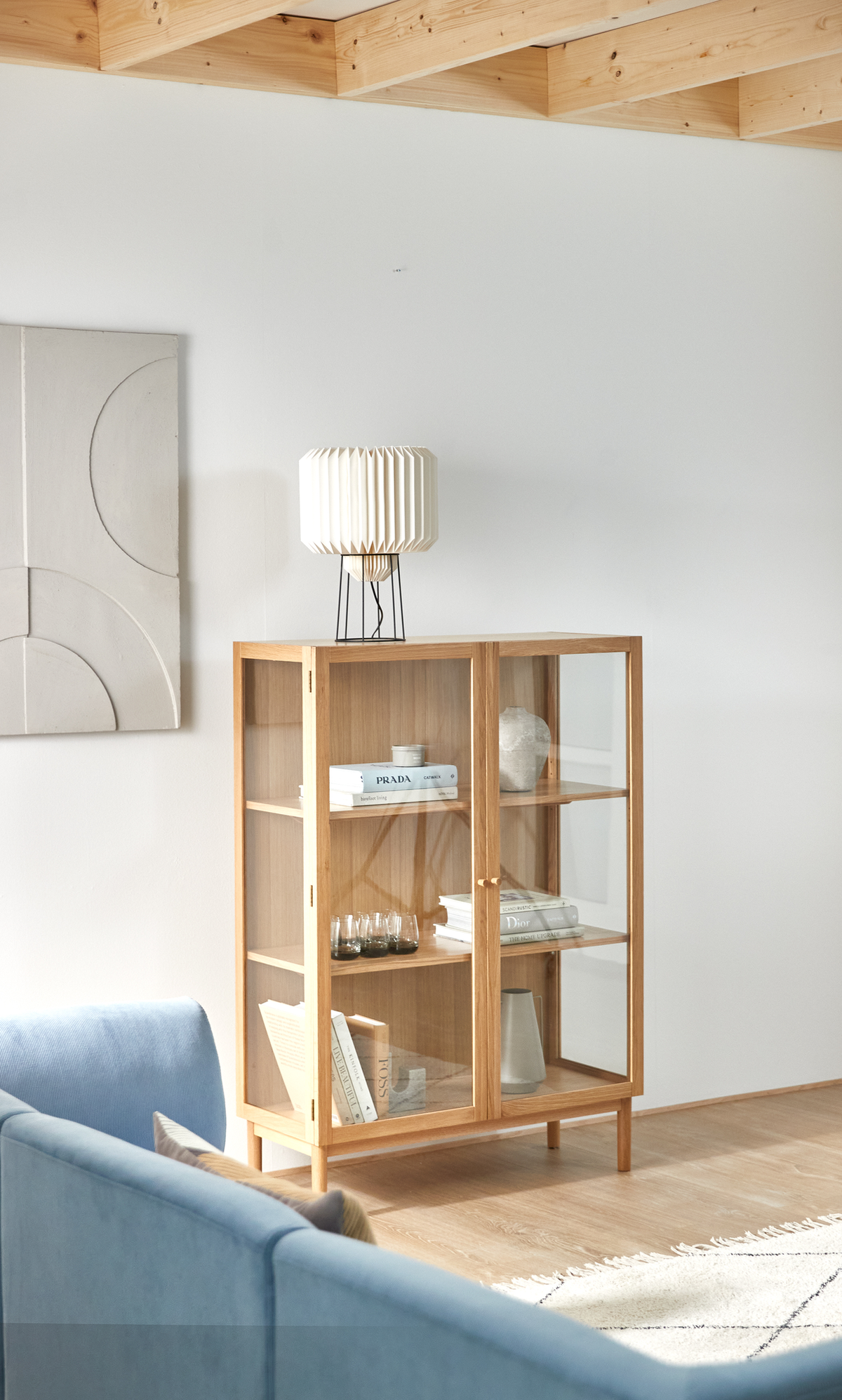 Lyst skandinavisk stue med vitrineskab i eg med glaslåger, stylet med bøger som Prada og Dior, små glas og keramik, med hvid plisseret bordlampe ovenpå, blå sofa og lys tæppe.