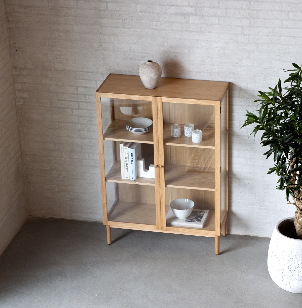 Vitrineskab i lys eg med glaslåger, stylet med tallerkener, skåle, små lys og bøger – herunder en med titlen PRADA – i et minimalistisk interiør med potteplante.