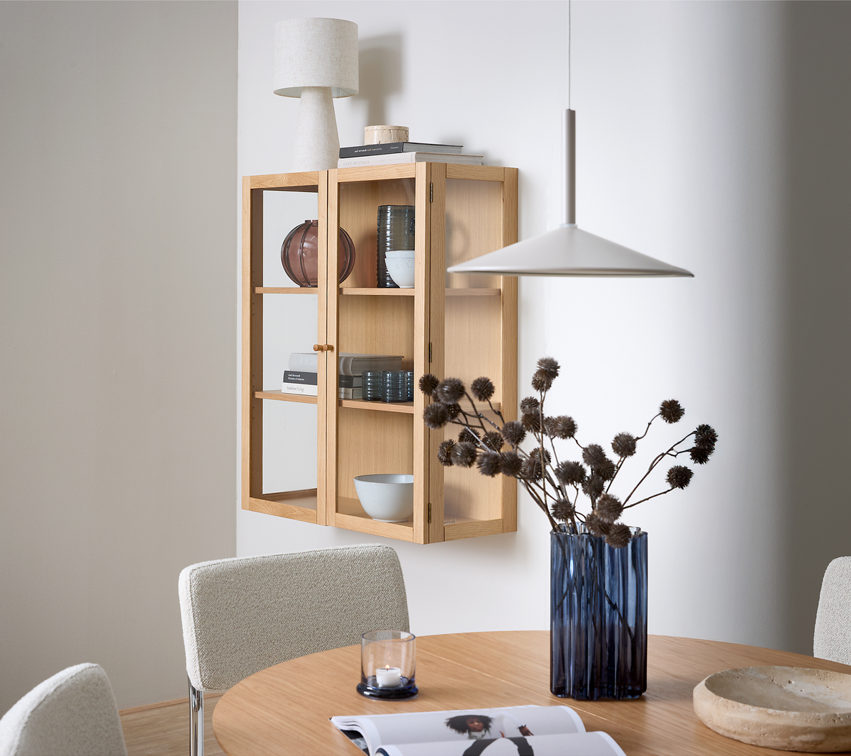 Spiseplads med rundt træbord, blå glasvase med tørrede grene, væghængt vitrineskab i eg med bordlampe, bøger og keramik samt en hvid pendel over bordet.