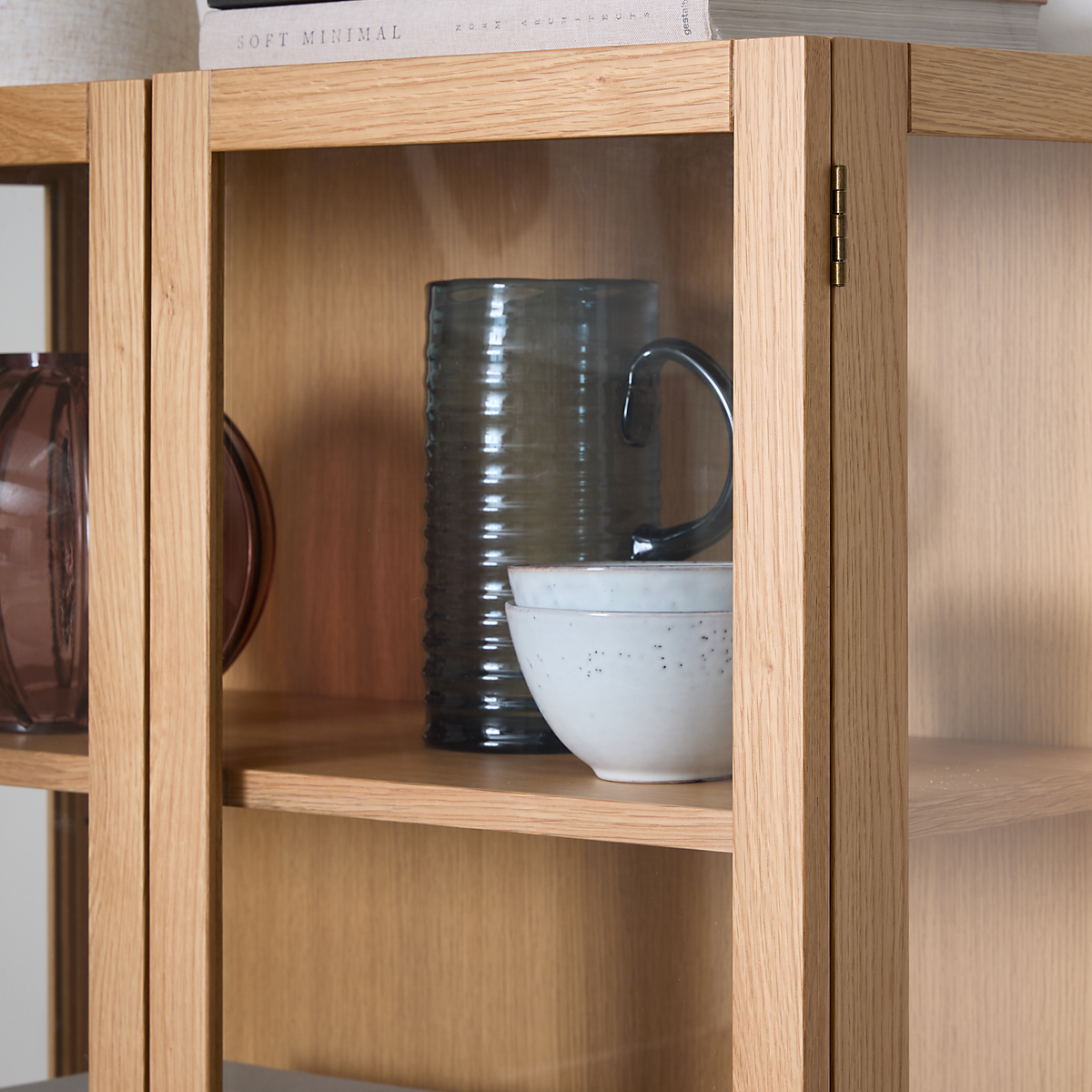 Nærbillede af et vitrineskab i egetræ med glasdør, en rillet glas-kande og to hvide skåle på hylden samt en bog med titlen Soft Minimal ovenpå.