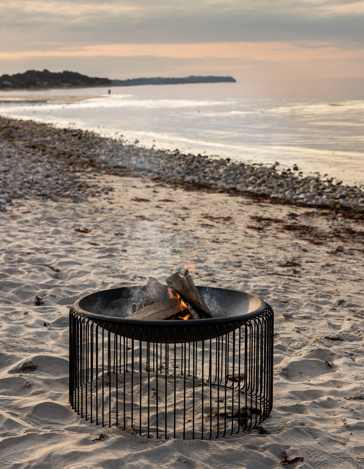 Moderne sort bålsted på sandstrand med brænde, hav i baggrunden ved solnedgang.
