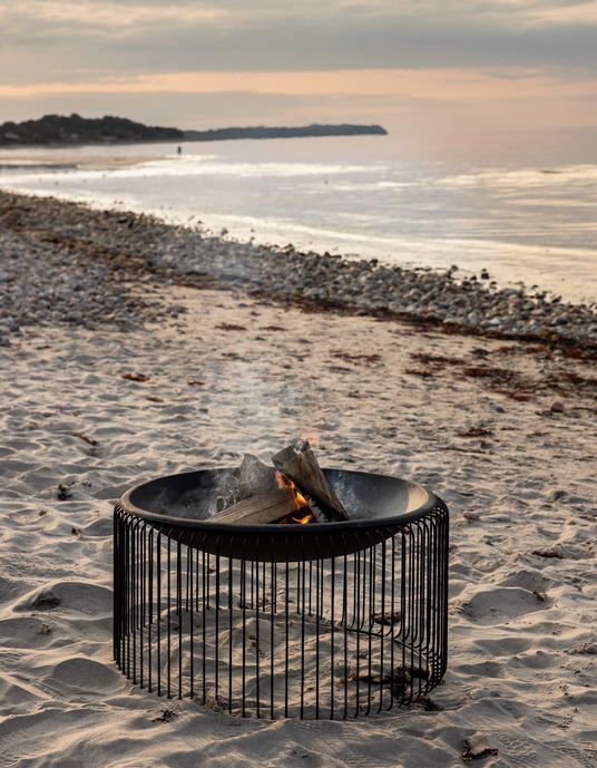 Moderne sort bålsted på sandstrand med brænde, hav i baggrunden ved solnedgang.