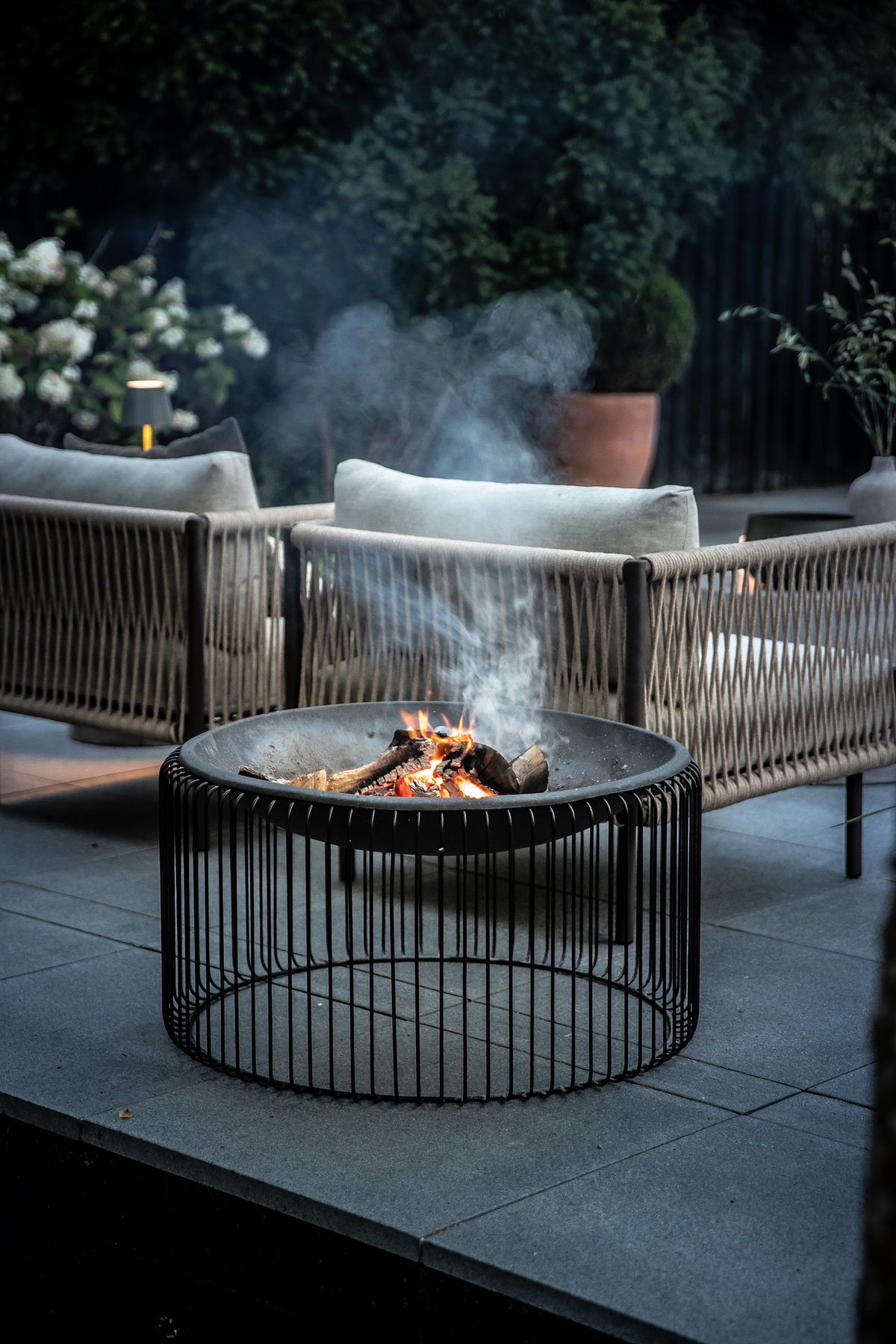 Moderne bålsted i sort metal med brænde, omgivet af flettede loungestole og krukplanter på en terrasse i skumringen.