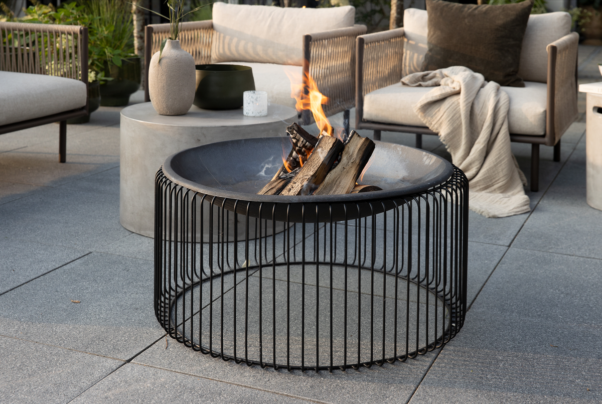 Moderne bålsted i metal med brænde på terrasse med loungemøbler, betonbord og plaid.