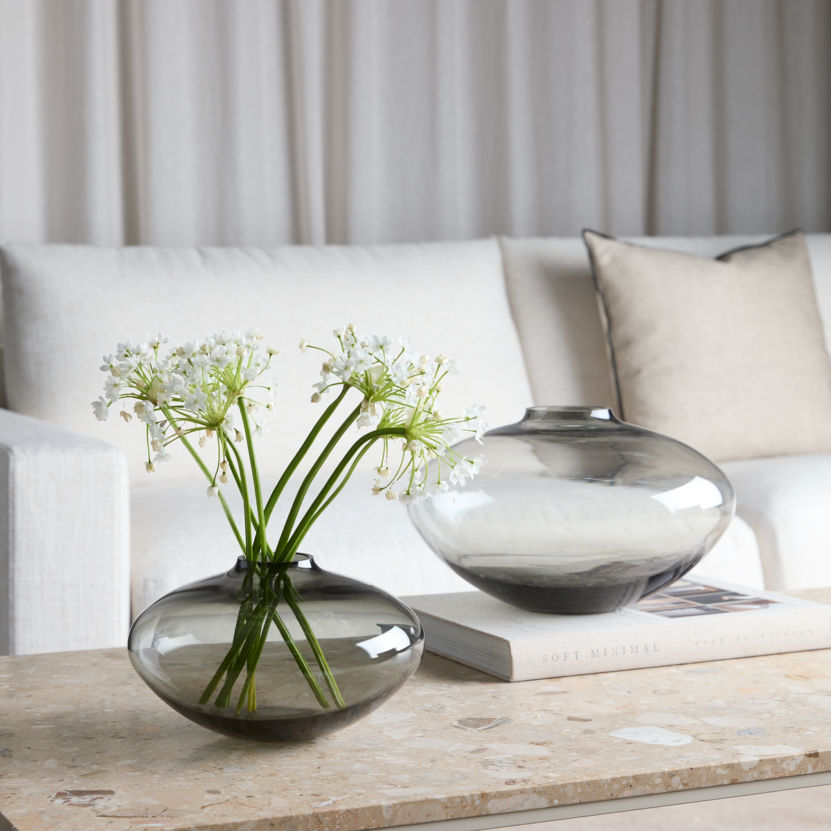 To røggrå glasvaser på et stenbord foran en lys sofa; den forreste med hvide blomster, den bagerste står på bogen “Soft Minimal”.