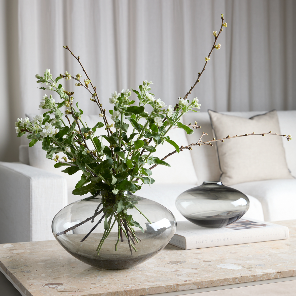 Rund røgfarvet glasvase med grønne grene og hvide blomster på et lyst stenbord i en lys stue; en anden lav glasvase står på en bog med titlen Soft Minimal, med sofa, lyse puder og gardiner i baggrunden.