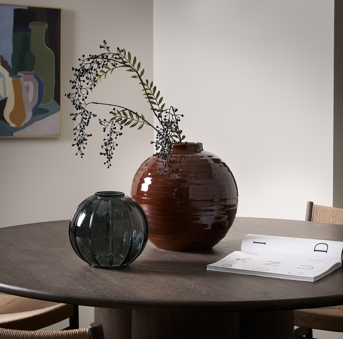Rundt spisebord i mørkt træ med en stor brun keramikvase med gren, en mindre grøn glasvase og en opslået bog med “PAI” og “GUID”; abstrakt stillebenmaleri på væggen.