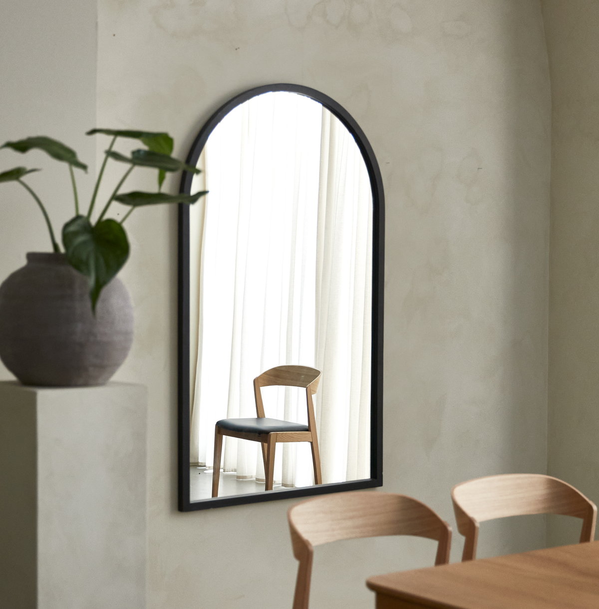 Bueformet vægspejl med sort ram; spejlet reflekterer en træstol med sort sæde foran lyse gardiner. I forgrunden ses træstole, et bord og en vase med grøn plante i et lyst, minimalistisk rum.