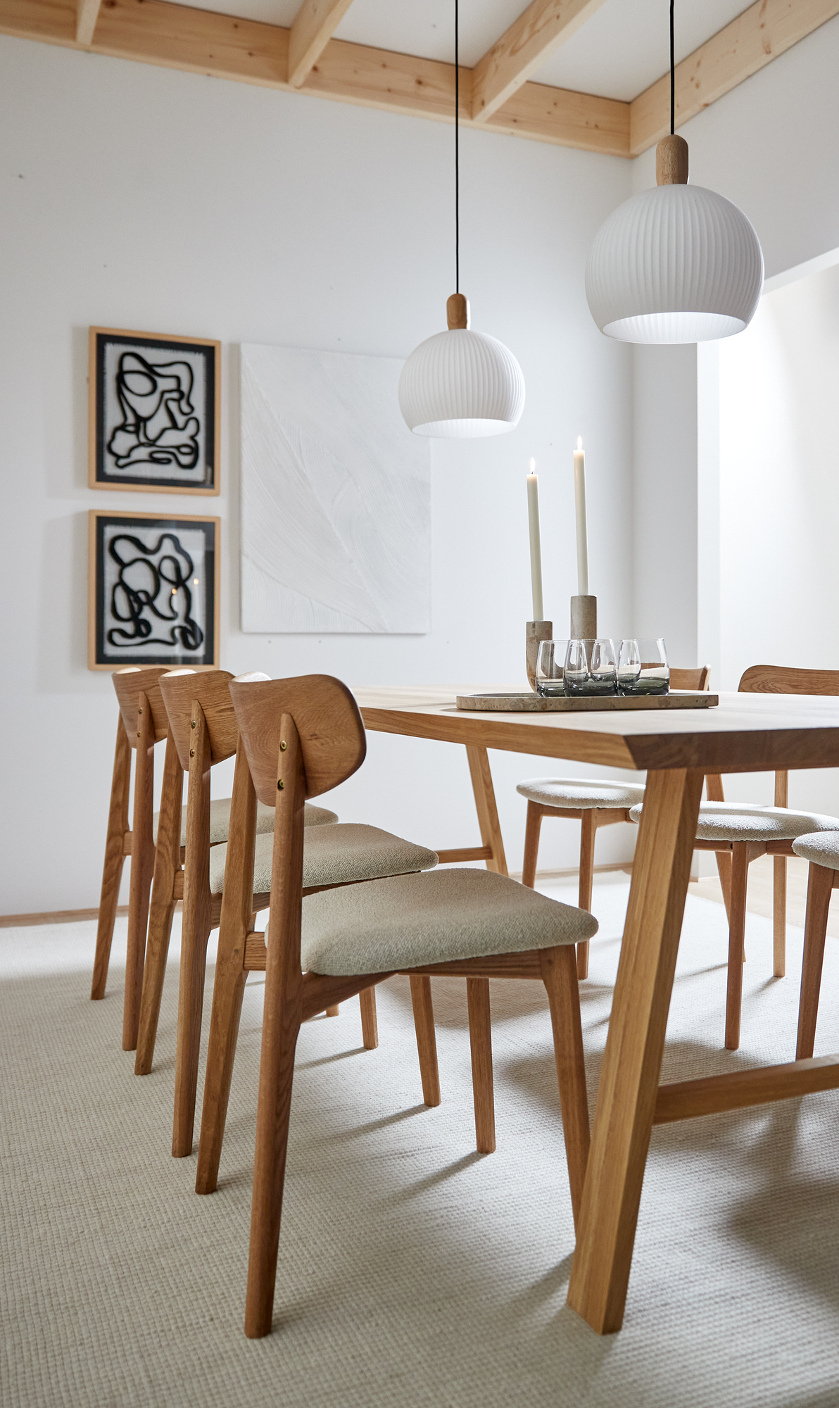 Lyst spiseområde med egetræsbord og seks polstrede stole, to hvide pendler over bordet, lys og glas på bakke samt abstrakt vægkunst på hvid væg.