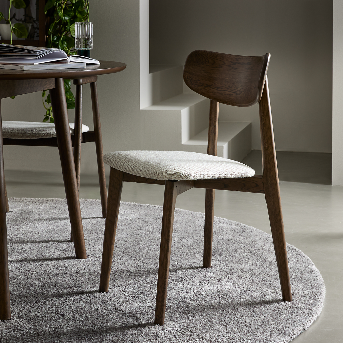 Moderne spisebordsstol i mørkt træ med lys polstret sæde på et rundt gråt tæppe ved siden af et rundt bord i et minimalistisk spiseområde.