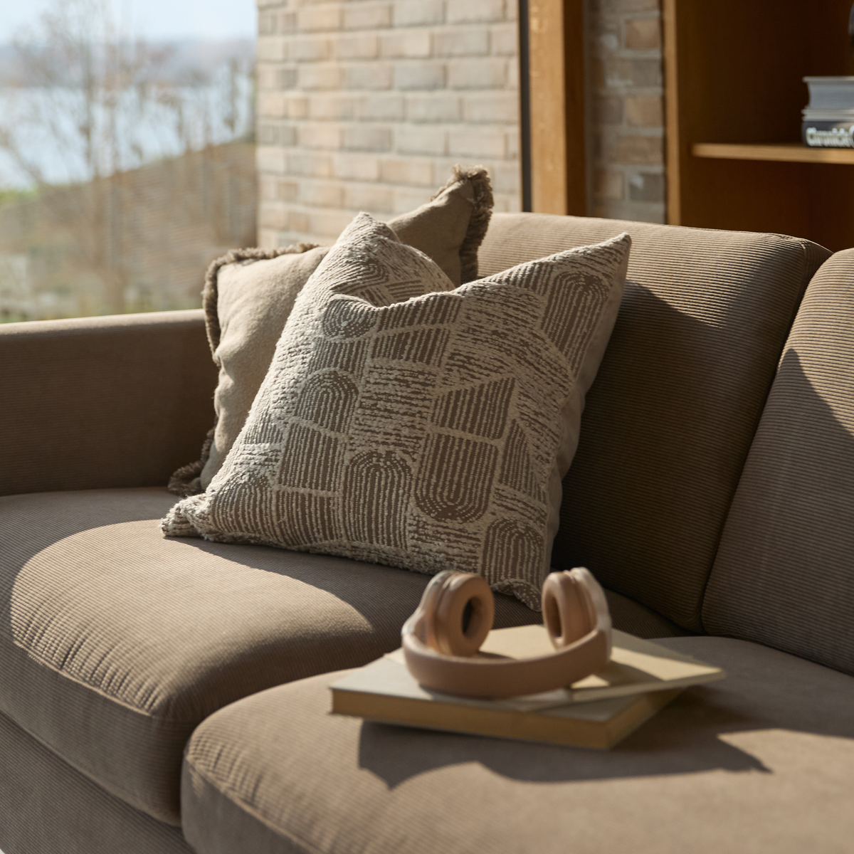 Beige sofa med mønstrede puder, en bog og beige hovedtelefoner i en solrig stue.