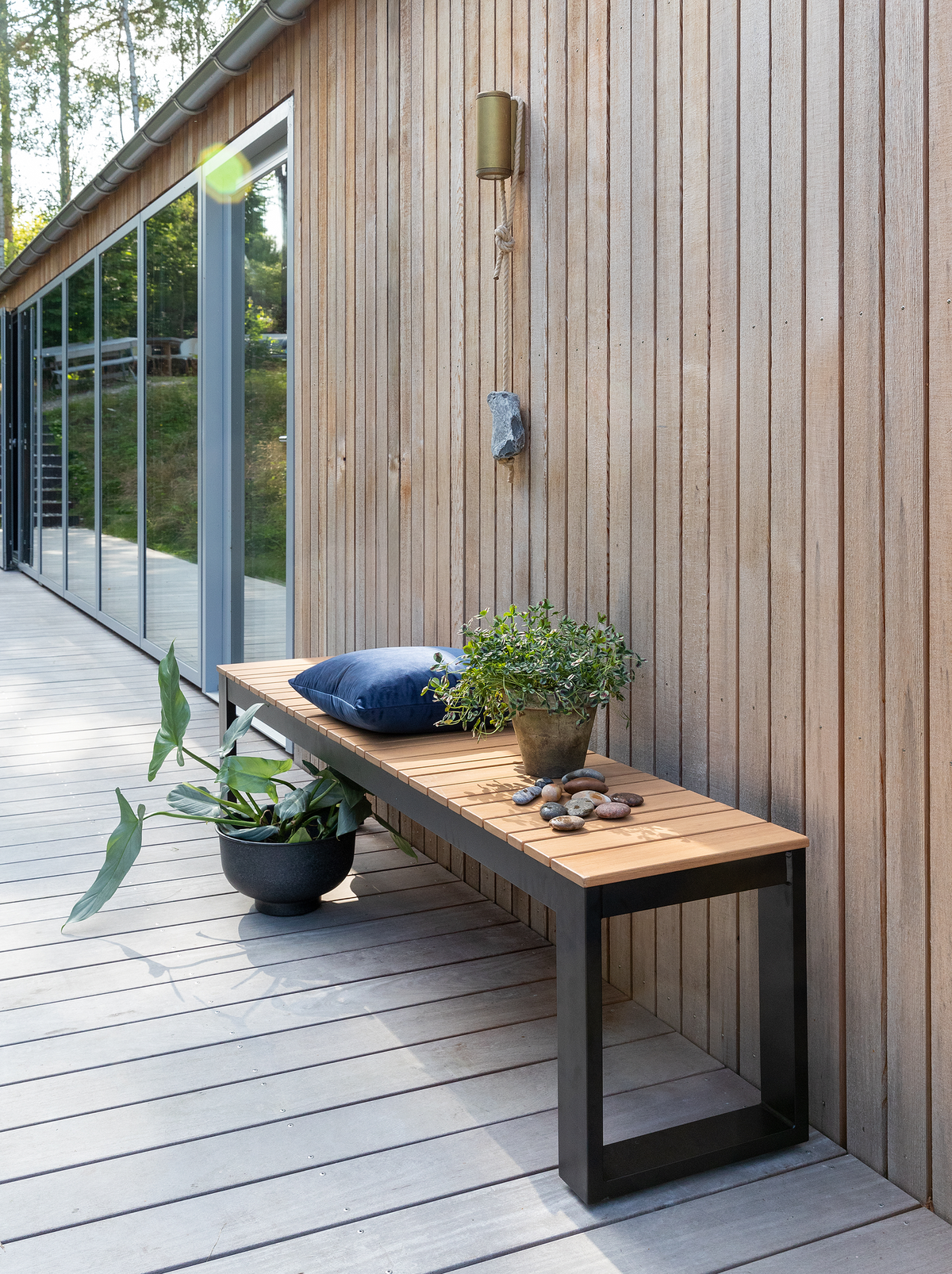 Træterrasse langs en træbeklædt husfacade med bænk i træ og sort metal, blå pude, potteplanter og sten i dagslys.