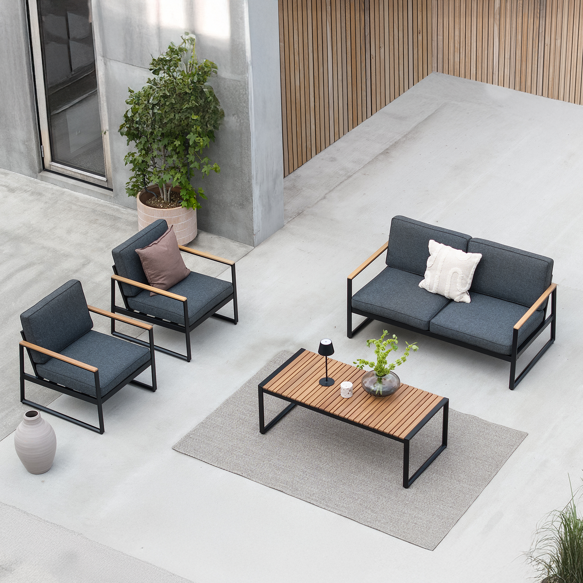 Moderne udendørs loungesæt med to lænestole og en to-personers sofa i mørkegråt med sorte stel og træarmlæn, rektangulært sofabord i lameltræ på gråt tæppe, krukkeplante, lille bordlampe og vase; dagslys i gårdrum.