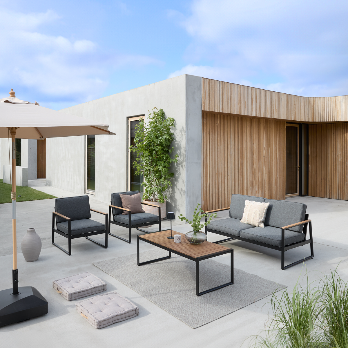 Moderne terrasse med gråt loungesæt, to stole og bord i træ/metal på tæppe, parasol og krukkeplanter foran et hus med beton og træbeklædning under blå himmel.