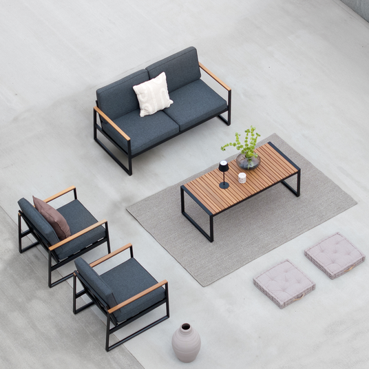 Moderne udendørs lounge med grå sofa og lænestole med træarmlæn, rektangulært træ/metal-bord på tæppe, lille bordlampe og vase med grønne grene, to siddepuder på gulvet og en stor gulvvase.