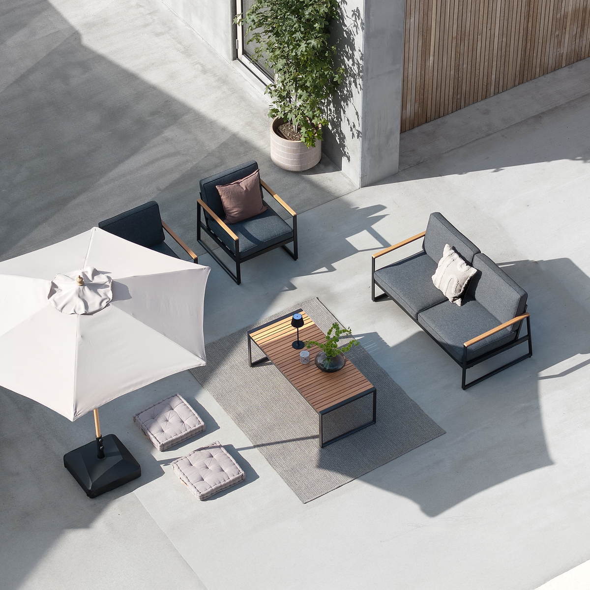 Moderne udendørs lounge med grå sofa og stole, træbord på tæppe, parasol og potteplante på betonterrasse, set oppefra.